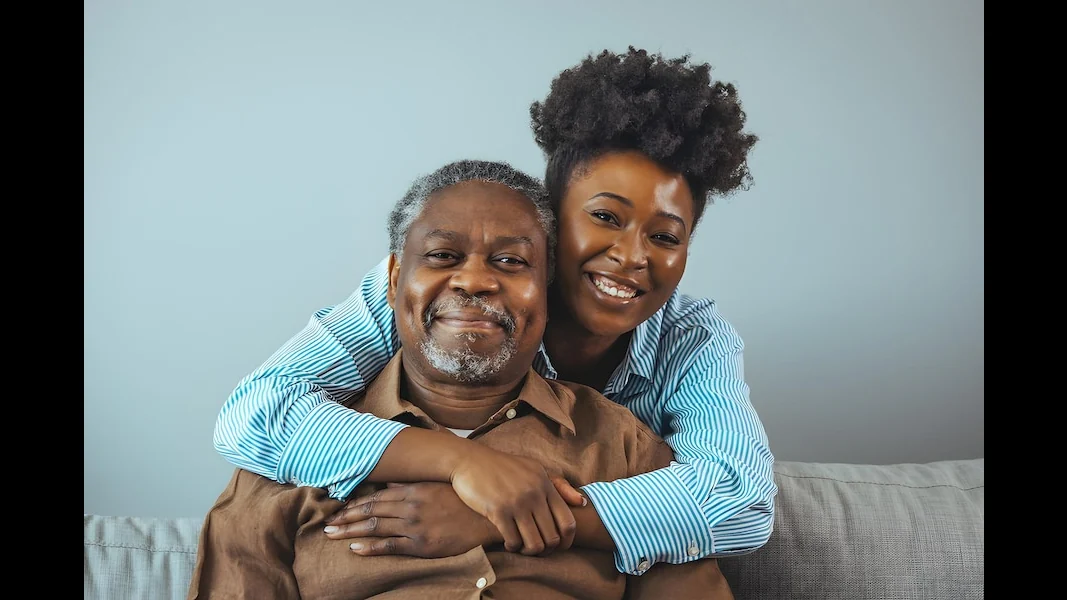 An adult daughter holds her elderly father close on the sofa at home, the kind of moment continuous care protects.